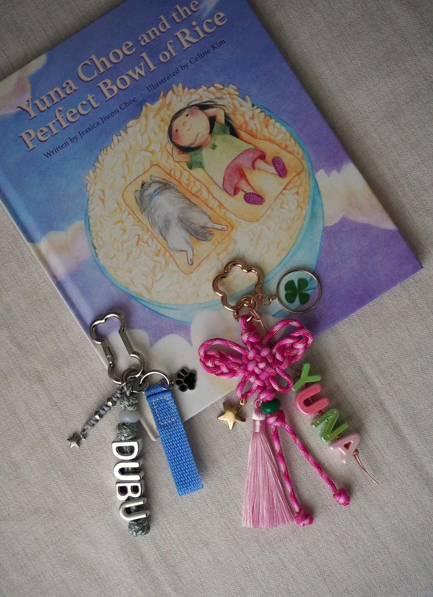 Two commissioned maedeup keychains side by side on linen — a silver DUBU keychain with woven knots and letter beads, and a pink butterfly knot keychain with YUNA letter beads, gold star charm, and tassel — resting on a children's book