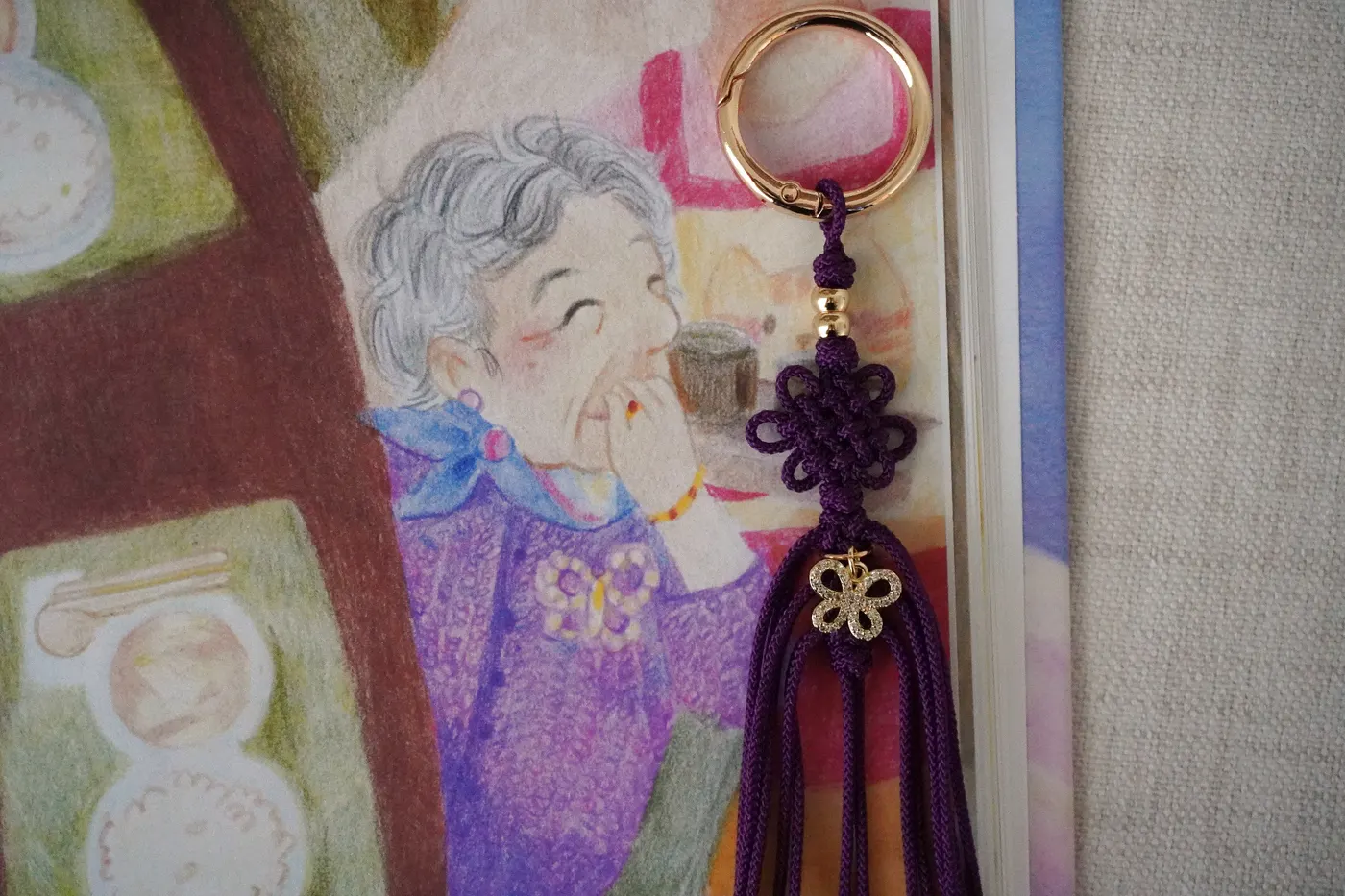 Deep purple commissioned maedeup norigae keychain with a hand-tied knot, gold butterfly charm, and long tassel resting beside a children's book illustration of a smiling Korean grandmother.