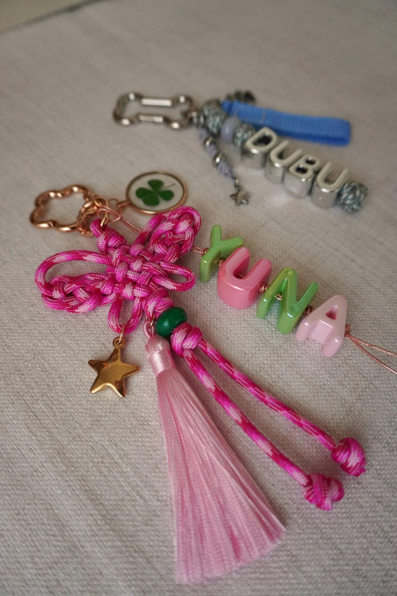 Two completed commissioned maedeup keychains side by side on linen — a pink butterfly knot with YUNA letter beads, gold star charm, and pressed clover, paired with a silver-and-blue DUBU keychain featuring woven knots and letter beads.