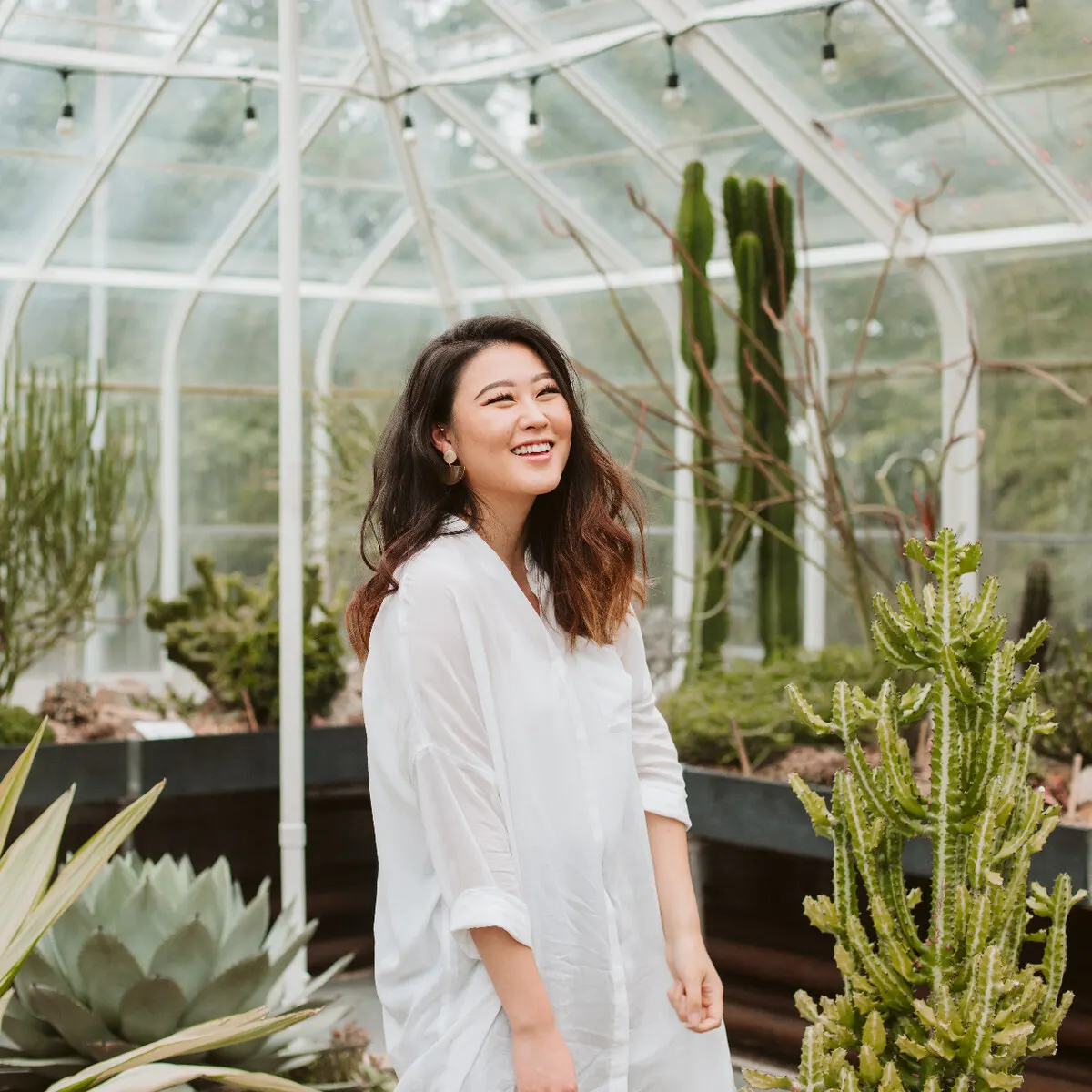 Staela Kim smiling inside a sunlit greenhouse surrounded by cacti and succulents